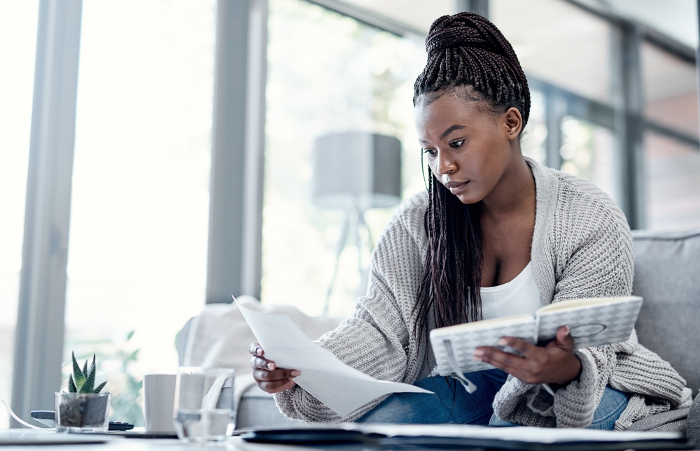 A woman sitting on a sofa at home, holding a notebook and reading a document with a focused expression, surrounded by natural light from large windows