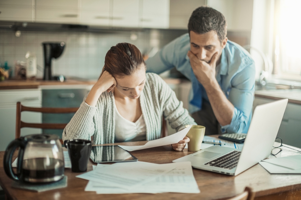 A couple sitting at a kitchen table reviewing bills and financial documents, looking concerned, with a laptop, coffee pot, and paperwork spread out in front of them.