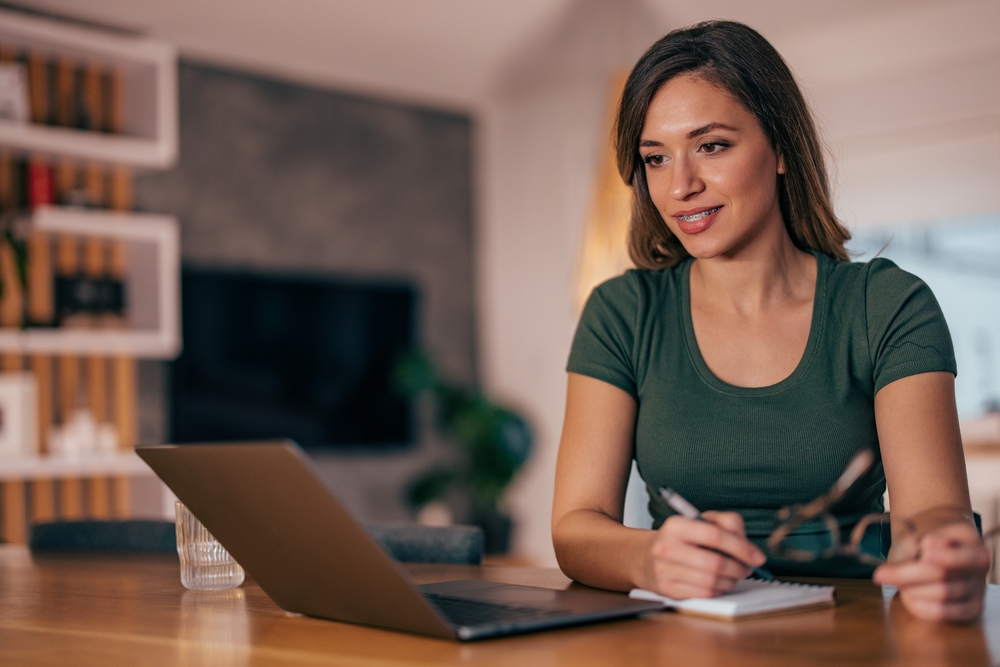 A young woman with shoulder-length brown hair, wearing a green t-shirt, sits at a wooden table and looks at a laptop screen. She is holding a pair of glasses in one hand and a pen in the other, writing in a small notebook. The background is a modern, blurred living space with shelves, a television screen, and soft indoor lightin