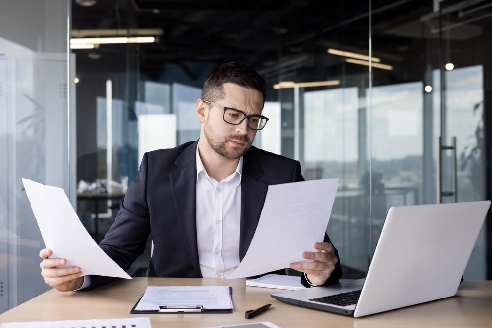 A focused professional man wearing glasses and a dark suit sits at a light wood desk in a modern office. He is intently reviewing and comparing two white paper documents, holding one in each hand. A white laptop, a clipboard with papers, and a pen are also on the desk in front of him. Glass walls and a blurred office background are visible behind him.